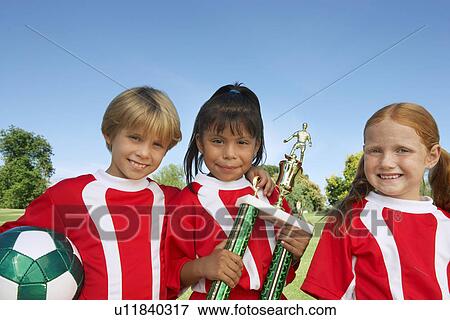 Stock Photo - Three children (7-9 years) holding soccer ball and trophy on soccer field, portrait. Fotosearch