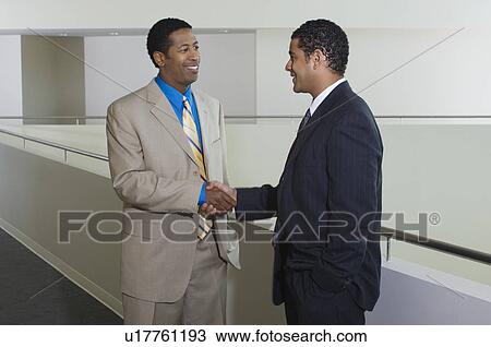 Stock Image - Two business men shaking hands in office hallway. Fotosearch