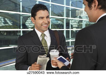 Stock Image - Two businessmen with take away coffee, newspaper, and PDA, outdoors. Fotosearch