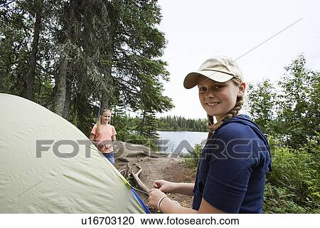 Stock Image - Two teenage girls standing by tent near lake, portrait. Fotosearch