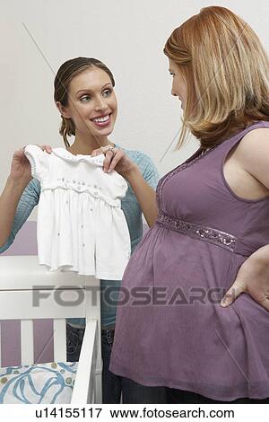 Stock Photo - Two women looking at baby clothes by cradle. Fotosearch