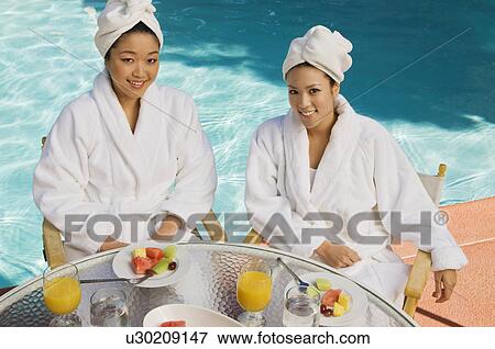 Stock Photo - Two young women wearing bathrobes by swimming pool, portrait. Fotosearch