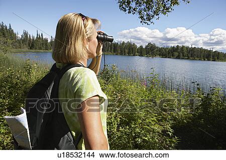 Picture - USA, Alaska, woman looking through binoculars at lake. Fotosearch