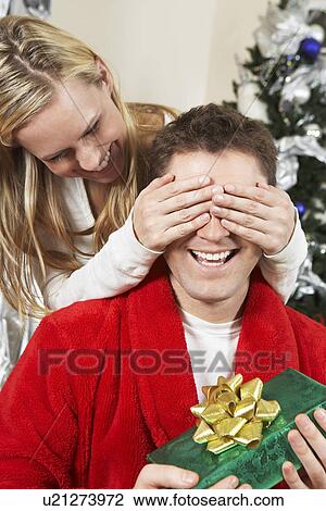Stock Image - Woman covering eyes of man holding present by Christmas tree. Fotosearch