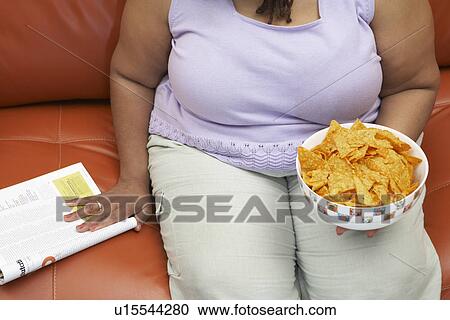 Stock Image - Woman sitting on couch with magazine and crisps, mid section. Fotosearch