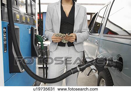 Stock Image - Woman standing by van with fuel pump counting money, mid section. Fotosearch