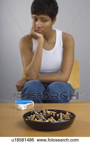 Stock Photograph - Woman staring at full ashtray of cigarettes on table. Fotosearch