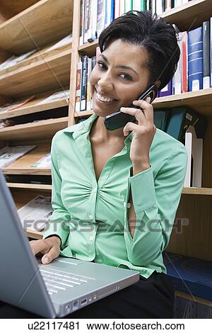 Stock Image - Woman using laptop and mobile phone in library. Fotosearch