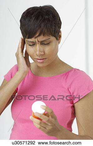 Stock Image - Woman with hand on head, holding pill bottle indoors. Fotosearch
