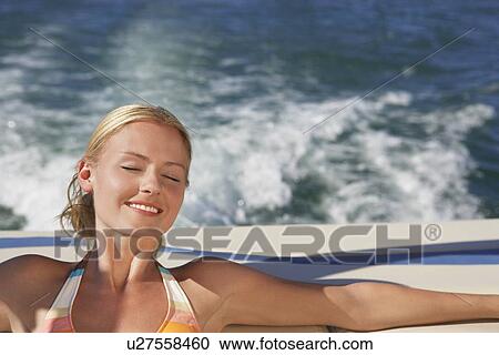 Stock Image - Young woman with eyes closed relaxing on boat, smiling. Fotosearch