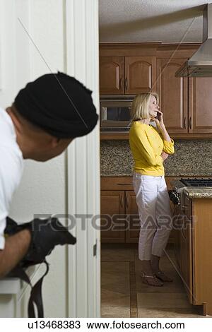 Stock Image - Burglar holding gun approaches woman on phone in kitchen. Fotosearch