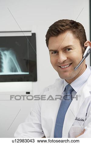 Stock Image - Doctor wearing earpiece in hospital, portrait. Fotosearch
