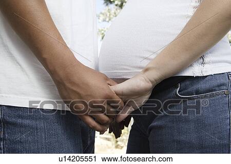 Stock Image - Expectant couple holding hands, mid section. Fotosearch