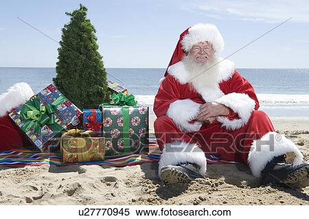 Stock Photography - Father Christmas sits on the beach with a tree and presents. Fotosearch