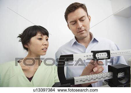 Man standing on weighing scales beside nurse in hospital View Large Photo Image Picture - Man standing on weighing scales beside nurse in hospital. Fotosearch