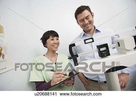 Stock Image - Man standing on weighing scales beside nurse in hospital. Fotosearch
