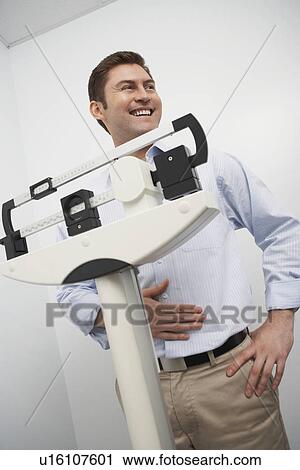 Stock Image - Man standing on weighing scales in hospital. Fotosearch