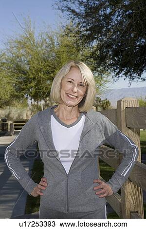 Stock Image - Mature adult woman stands smiling, in tracksuit with hands on hips. Fotosearch