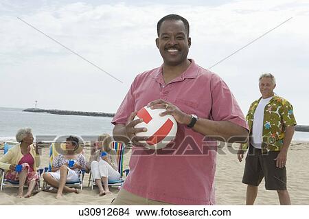 Picture - Mature man stands with volleyball on beach, friends grouped behind. Fotosearch