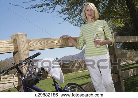 Stock Photograph - Mature woman stands with mountain bike at fence. Fotosearch