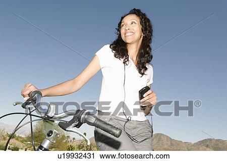 Stock Image - Mid adult woman listens to MP3 holding handlebars on mountainbike. Fotosearch