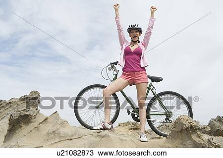 Stock Image - Mid adult woman stands with mountain bike and arms raised. Fotosearch