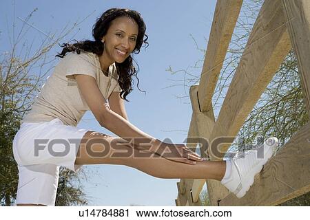 Stock Image - Mid adult woman stretches hamstring against fence. Fotosearch