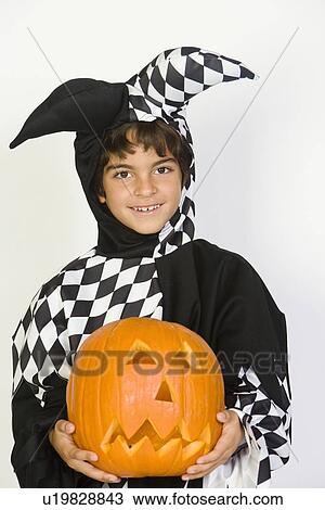 Stock Image - Portrait of boy (7-9) wearing jester costume, with jack-o-lantern. Fotosearch