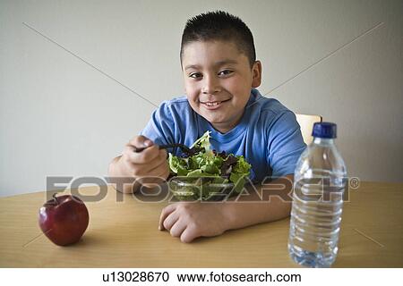 Stock Image - Pre-teen (10-12) boy sitting at desk and eating salad. Fotosearch