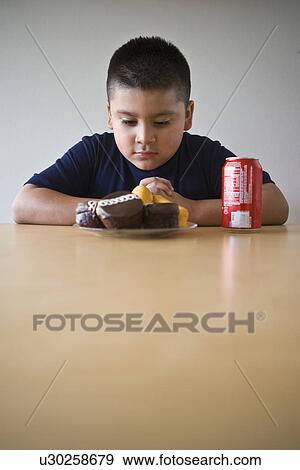 Stock Photo - Pre-teen (10-12) boy sitting at desk and looking at dessert. Fotosearch