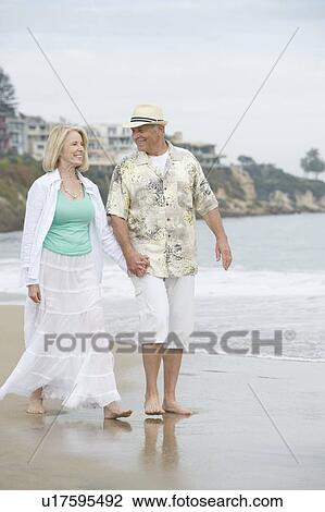 Stock Image - Senior couple walk holding hands on beach. Fotosearch