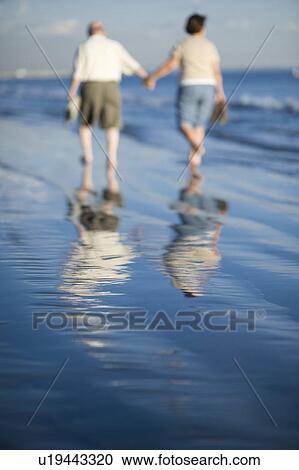 Stock Image - Senior couple walking on beach, reflecting in water. Fotosearch