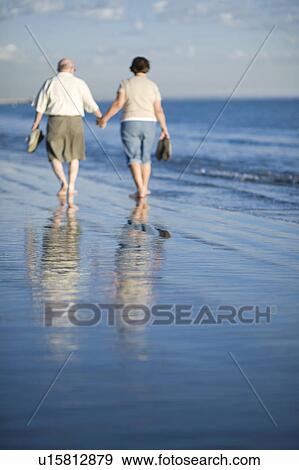 Stock Photo - Senior couple walking on beach, reflecting in water. Fotosearch