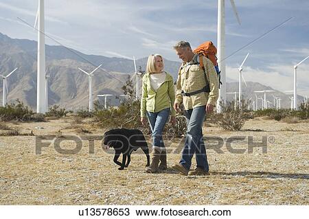 Stock Image - Senior couple walking with dog near wind farm. Fotosearch