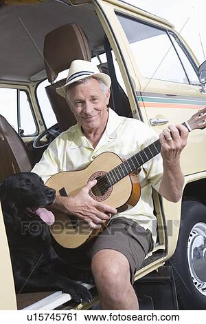 Stock Image - Senior man sits in his campervan with a guitar and his pet dog. Fotosearch