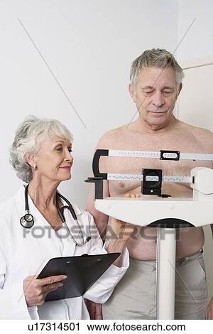 Stock Image - Senior medical practitioner stands with clip-board and client on measuring scales. Fotosearch