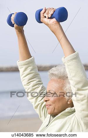 Stock Photography - Senior woman excercises with dumbbells on beach. Fotosearch