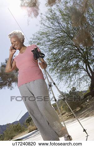 Stock Image - Senior woman stands on phone with walking poles. Fotosearch
