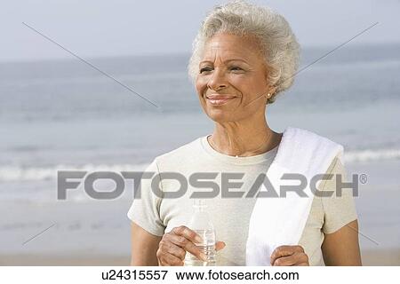 Senior woman stands with drinking water and towel over her shoulder on beach View Large Photo Image Stock Photo - Senior woman stands with drinking water and towel over her shoulder on beach. Fotosearch