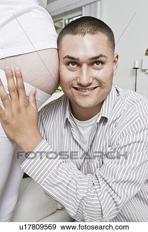 Stock Photo - Smiling man with head at pregnant woman's abdomen. Fotosearch