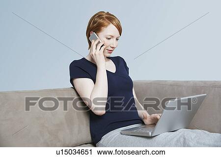 Stock Image - Woman using laptop and mobile phone on sofa. Fotosearch