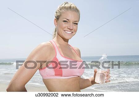 Stock Image - Young woman holding bottle of water at the beach. Fotosearch