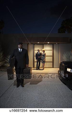 Picture - Chauffeur carries bags for couple standing at lit entrance. Fotosearch