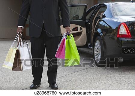 Stock Image - Chauffeur holds shopping bags in driveway near luxury vehicle. Fotosearch