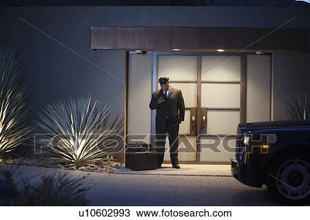 Stock Image - Chauffeur stands at lit entrance doorway with luggage. Fotosearch
