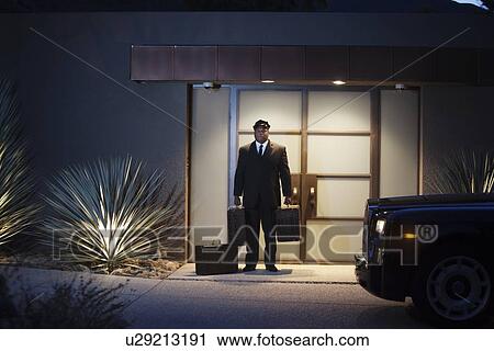 Stock Image - Chauffeur stands at lit entrance doorway with luggage. Fotosearch