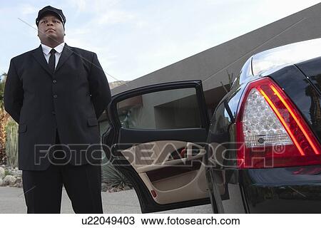 Stock Image - Chauffeur stands at open car door of luxury vehicle. Fotosearch
