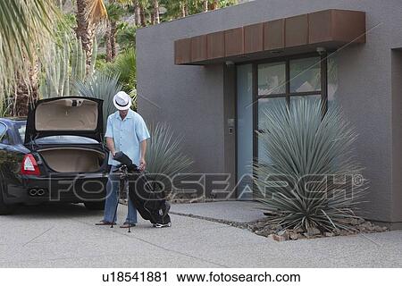 Stock Image - Man lifts golf bag into boot of luxury vehicle. Fotosearch