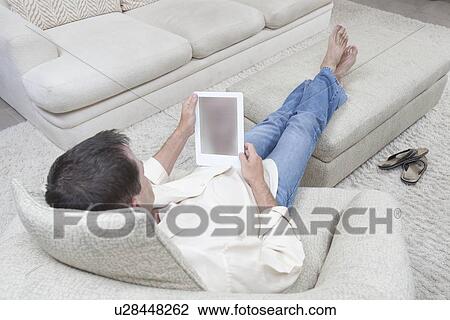 Stock Image - Man sits with feet up reading a digital book. Fotosearch