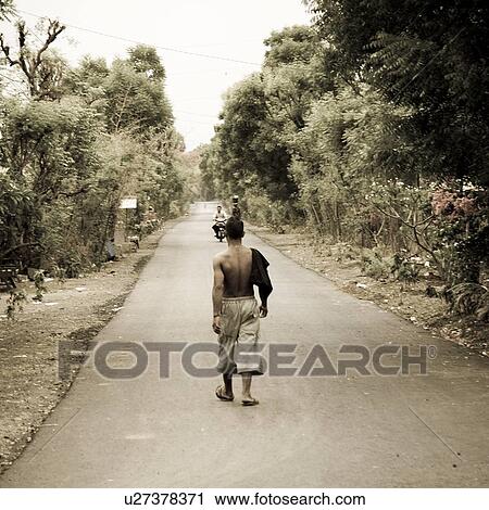 Stock Image - Back view of a man walking on roadway in Bali. Fotosearch